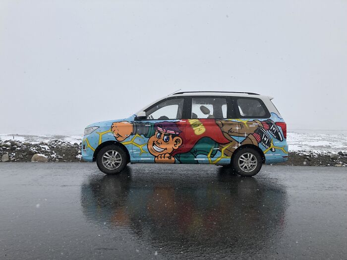 Car covered in colorful street art featuring a cartoon character, parked on wet pavement with snowy background.