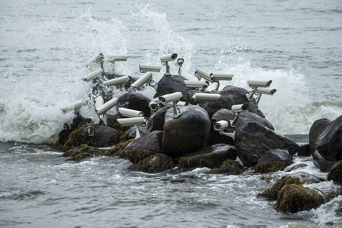 Surveillance cameras mounted on ocean rocks with waves crashing, a striking example of street art photos.