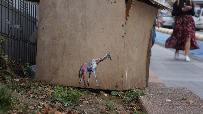 Small colorful street art of a giraffe painted on the side of a cardboard box near a sidewalk.