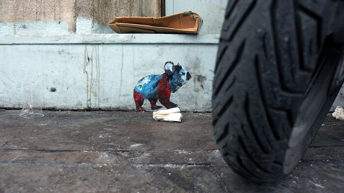 Colorful street art photo of a small bear painted on a wall near a large motorcycle tire on an urban sidewalk.