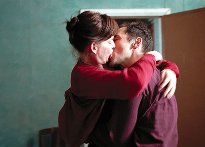 Two movie stars intimately embracing and kissing while filming love-making scenes on set in a dimly lit room.