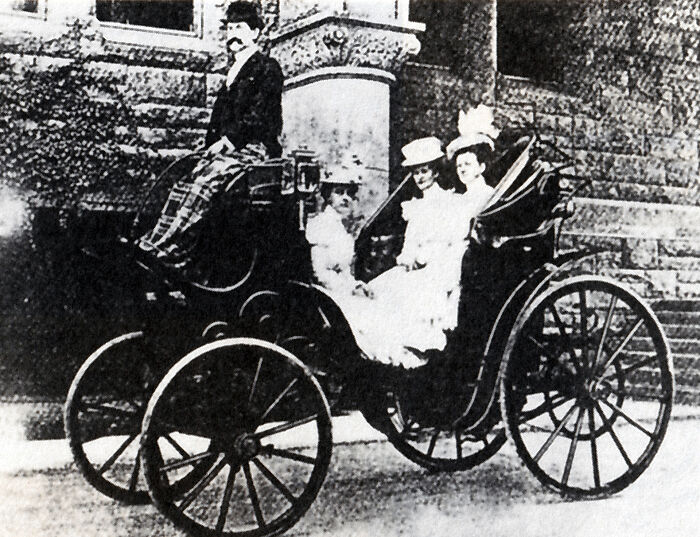 Early photo showing wild first cars with passengers dressed in vintage clothing seated in an antique vehicle.