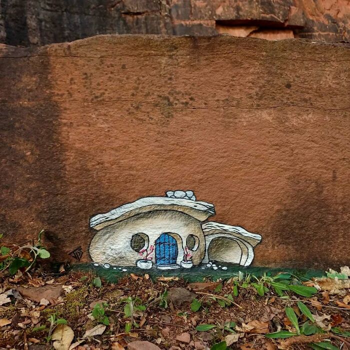 Street graffiti art of a small stone house interacting with natural surroundings on a brown rock surface.