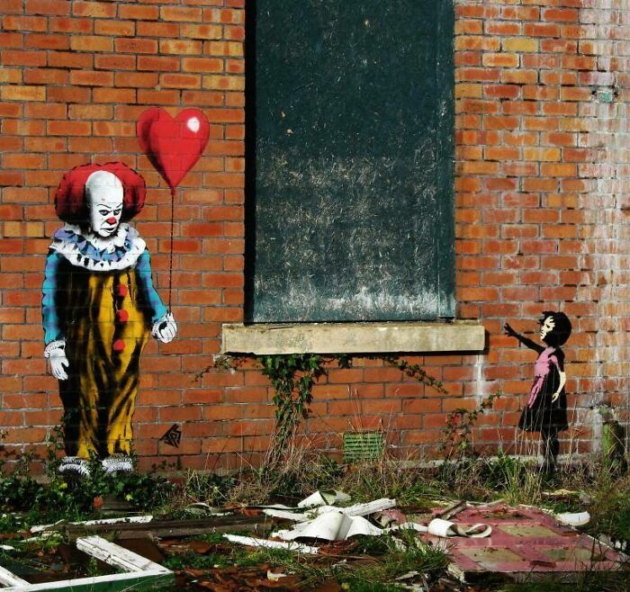 Graffiti art on brick wall shows clown holding heart balloon and a girl reaching out, creatively interacting with surroundings.