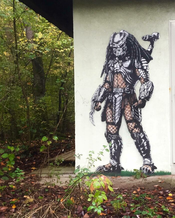 Graffiti of a Predator character interacting with the natural surroundings on a building wall near a forest.