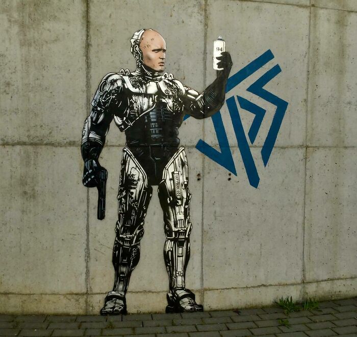 Graffiti of a cyborg holding a spray paint can, interacting creatively with a concrete wall and geometric shapes.