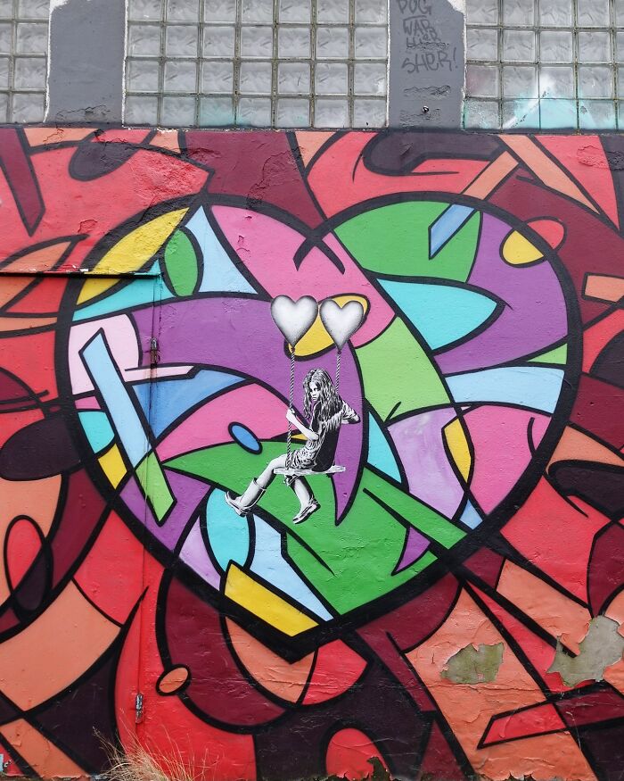 Colorful graffiti heart with a girl on a swing attached to heart-shaped balloons, showing interactive street art.