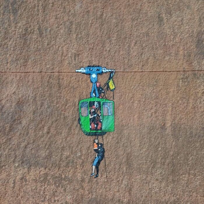 Street art graffiti on a wall showing a green cable car with people interacting, blending with the surroundings.
