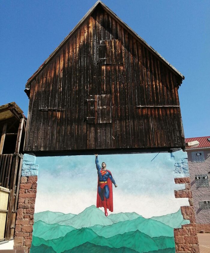 Street graffiti art interacting with surroundings featuring Superman flying above green mountains on a building wall.