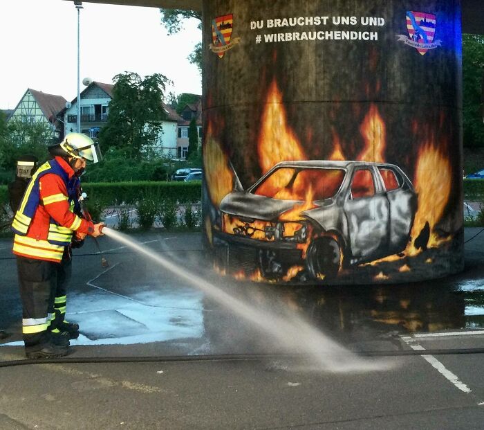 Firefighter spraying water at graffiti art of a burning car on a street structure, showcasing interactive street graffiti.