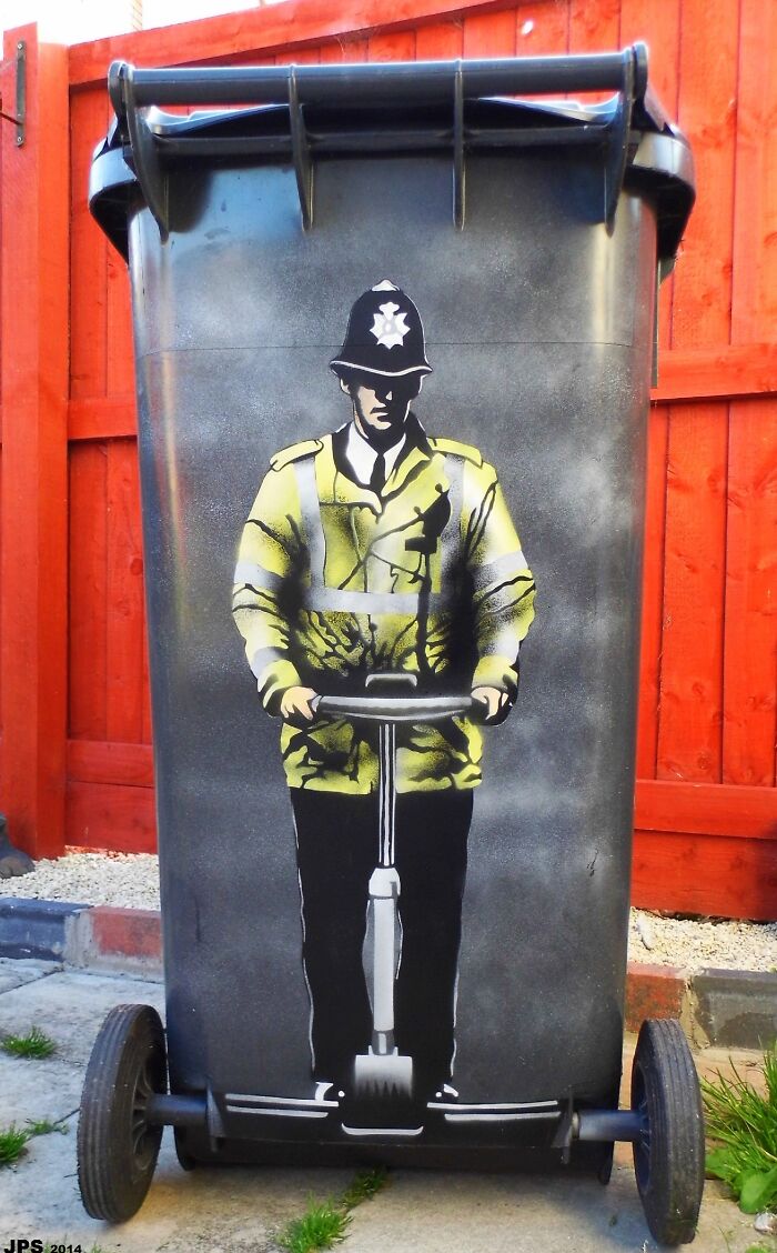 Street art of a police officer riding a Segway, painted on a black trash bin against a red wooden fence.