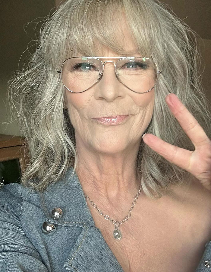 Jamie Lee Curtis close-up selfie wearing glasses and a denim jacket making a peace sign in a casual setting.