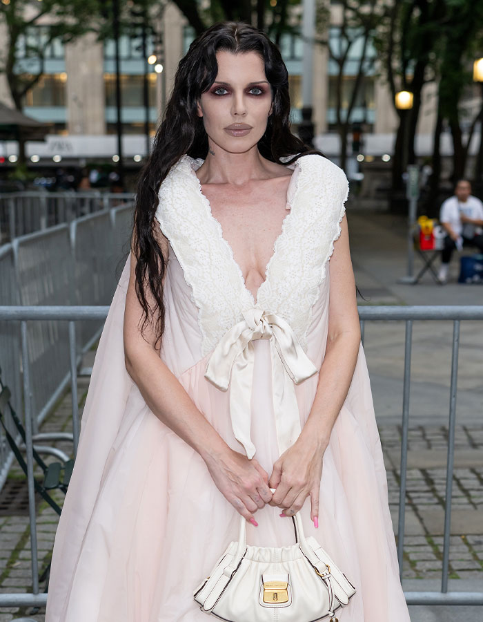 Julia Fox in a pale pink dress and carrying a white purse, posing outdoors with a serious expression about plastic surgeries.