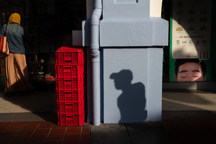 Shadow of a street photographer on a white wall beside red crates, capturing the beauty of ordinary life in a city setting.