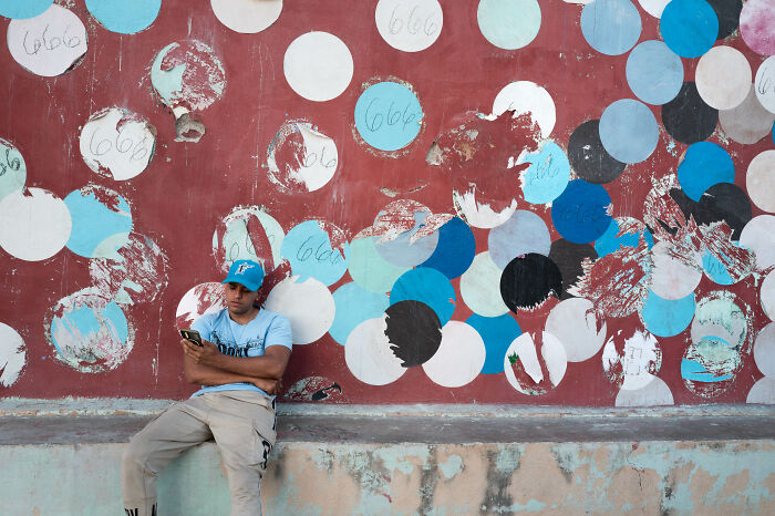 A street photographer captures a man sitting against a colorful wall with peeling paint and circular patterns.