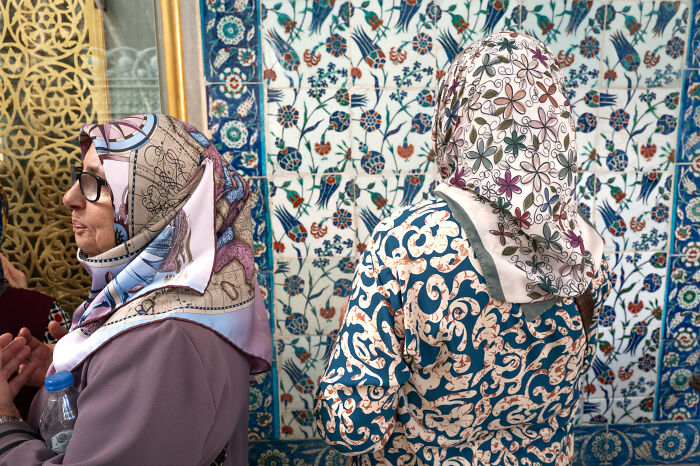 Two women wearing patterned headscarves stand in front of vibrant floral and geometric tiled walls, showcasing street photography.