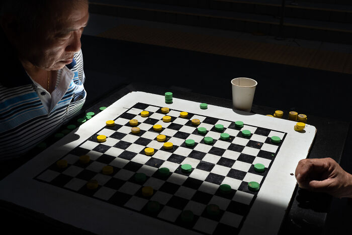 Street photographer captures the beauty of ordinary life showing two people playing checkers under soft light.