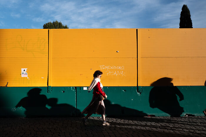 Street photographer captures woman walking past colorful wall with dramatic shadows, highlighting beauty of ordinary life.