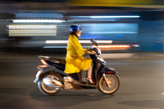 Woman in a yellow dress riding a scooter at night, captured by street photographer showcasing beauty of ordinary life.