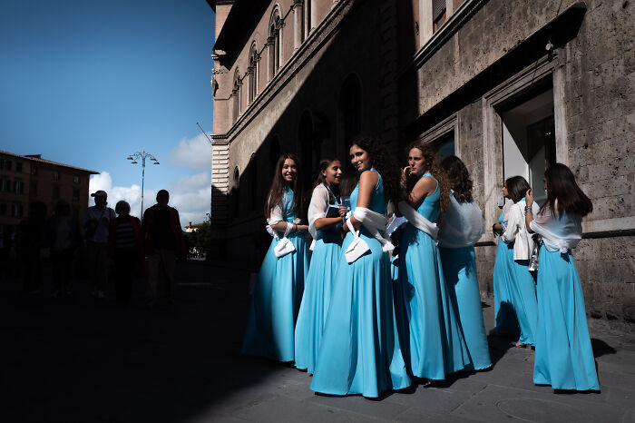 A street photographer captures a group of women in blue dresses gathering on a sunlit urban street, showcasing ordinary life.