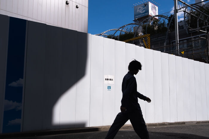 Silhouette of a person walking past a white wall under bright sunlight, showcasing street photography capturing ordinary life.