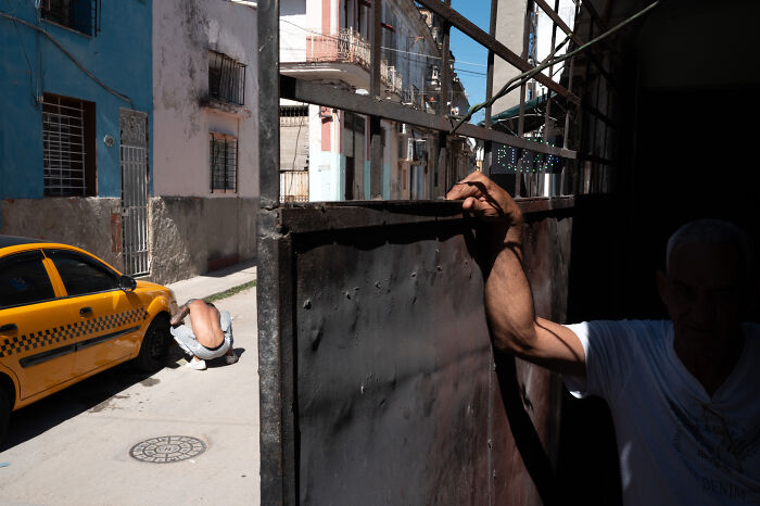 Man holding gate in shadow with yellow taxi and worker fixing tire on urban street, showing beauty of ordinary life in street photography.