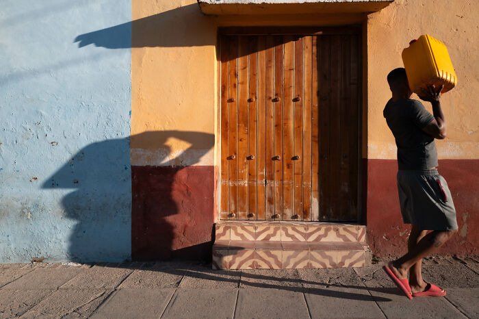Man carrying yellow container on shoulder walking past colorful wall, showcasing street photographer's capture of ordinary life.