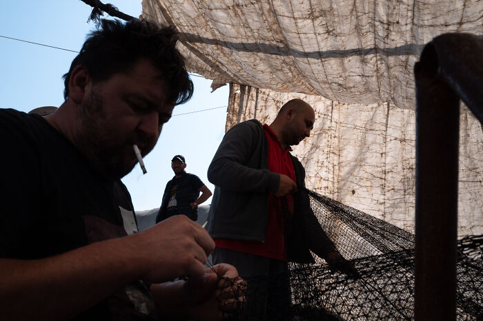 Two men repairing a fishing net under a tarp, captured by a street photographer showing ordinary life moments.