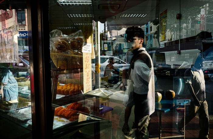 Reflection of pedestrians walking past a bakery, capturing ordinary moments in a poetic, cinematic style by the photographer.