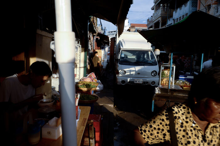 Cinematic image of a narrow street with people, food stalls, and a white van in warm, poetic light.