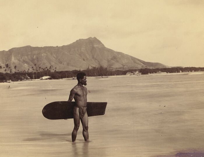 A historical photo of a shirtless man holding a wooden surfboard in shallow water with mountains in the background.