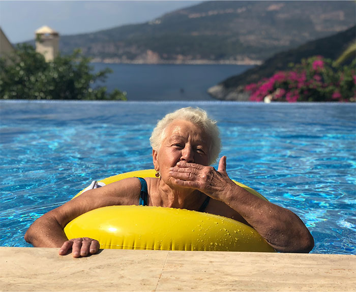Elderly woman in a pool with a yellow float, surrounded by scenic mountains and bright flowers on a sunny day.