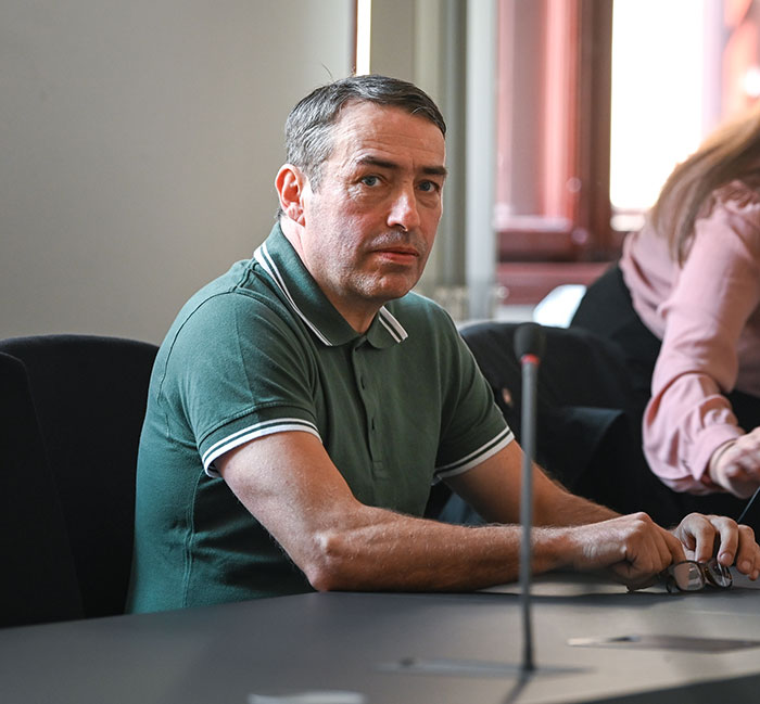 Man in a green polo shirt sitting at a table during a legal proceeding about radical right-winger gender change.