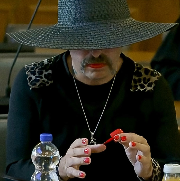 Person with mustache wearing a large hat, applying red nail polish, relating to radical right-winger gender change after conviction.