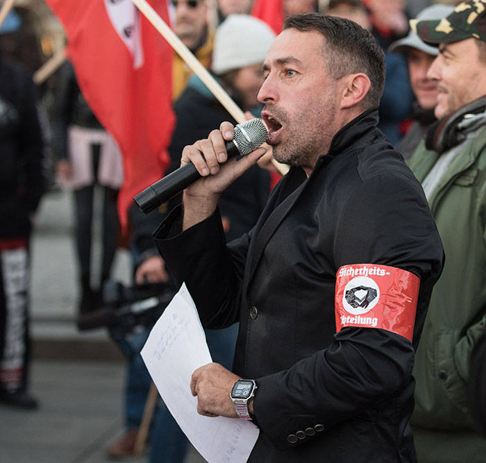 Man with red armband speaking into microphone at a public rally, related to radical right-winger gender change after conviction.