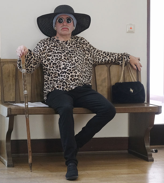Person wearing leopard print shirt, wide black hat, and sunglasses sitting with cane and handbag, radical right-winger gender change.