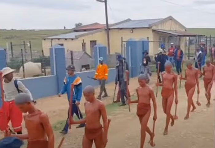 Boys walking in a mass circumcision ceremony supervised by adults in an outdoor rural area with buildings in the background - 2