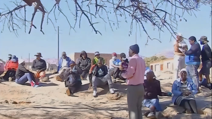 A group of people sitting and standing outdoors under a tree during a mass circumcision ceremony gathering. - 8