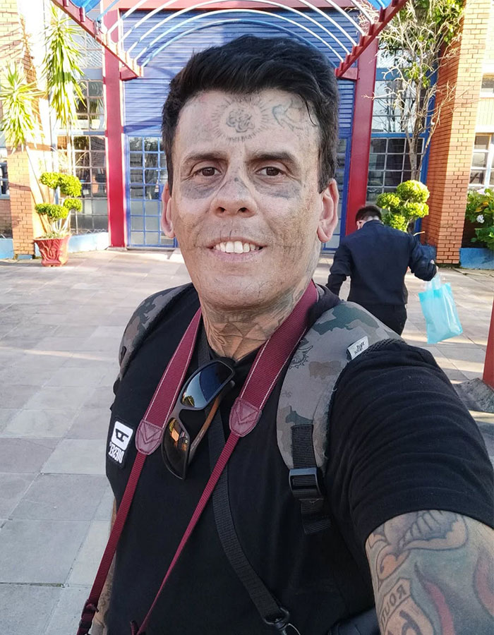 95% tattooed Brazilian Steve-O man showing his dramatic new look after tattoo removal outdoors with backpack and camera.
