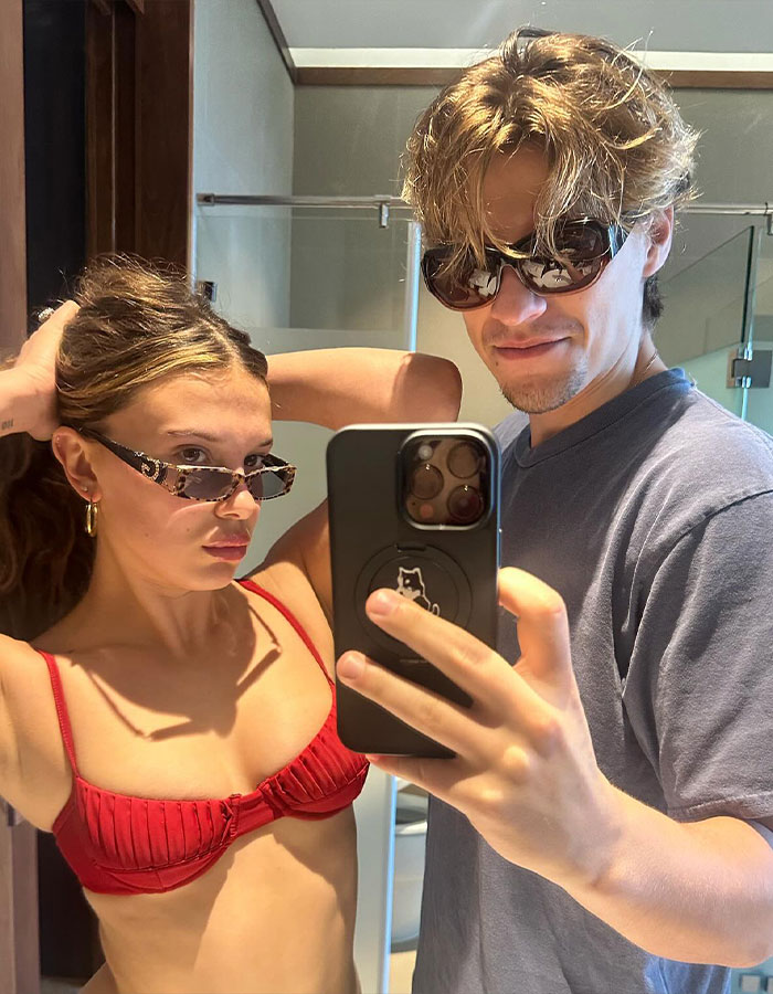 Young woman and man wearing sunglasses taking a mirror selfie, related to Millie Bobby Brown baby adoption debate.