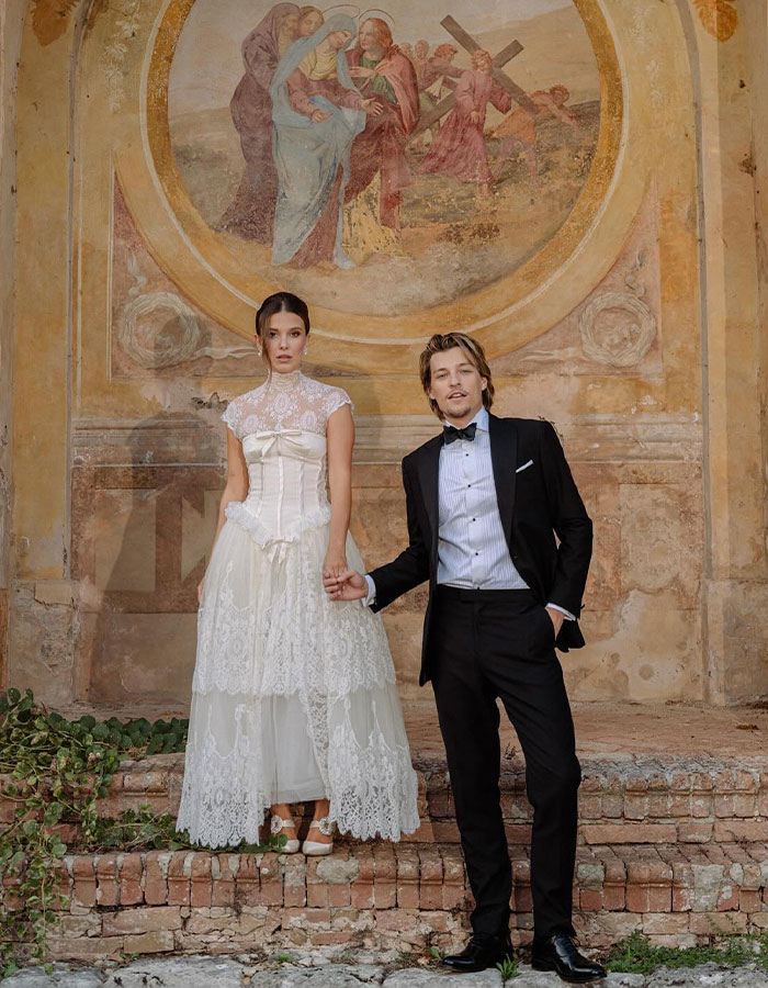 Young couple dressed formally standing in front of a historic wall painting, illustrating Millie Bobby Brown baby adoption debate.