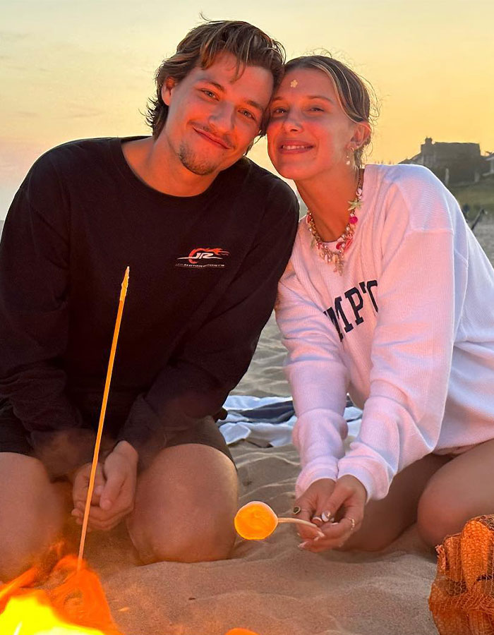 Millie Bobby Brown smiling with a friend on the beach at sunset, sparking baby adoption debate and fan reactions.