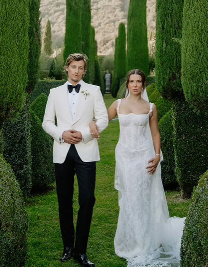 Millie Bobby Brown and Jake Bongiovi dressed formally walking together in a garden setting without their baby girl.