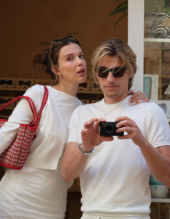 Millie Bobby Brown and Jake Bongiovi posing together indoors, capturing a casual selfie with camera in hand.
