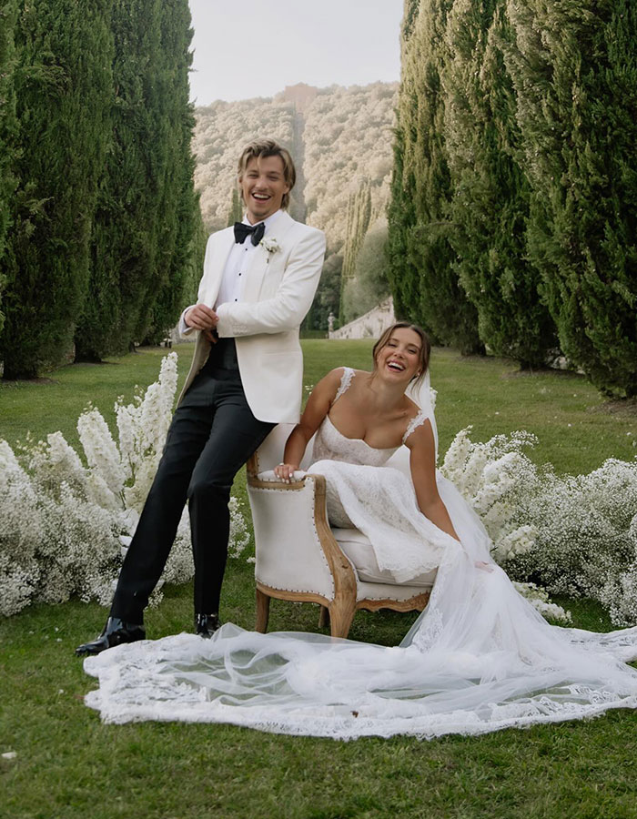Millie Bobby Brown and Jake Bongiovi smiling outdoors, dressed in wedding attire, surrounded by greenery and white flowers.