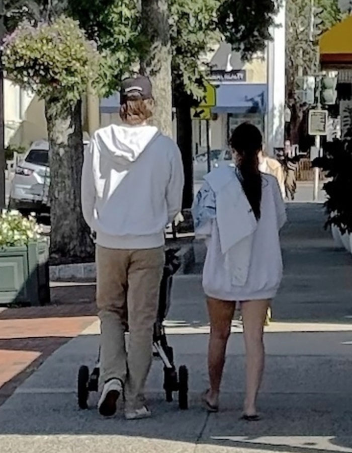 Millie Bobby Brown and Jake Bongiovi walking with baby stroller on a sunny day in a public outdoor setting.