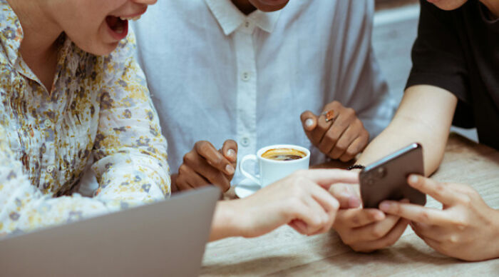 Three people gathered around a table debating a moral dilemma poll on a smartphone, with coffee and a laptop nearby.