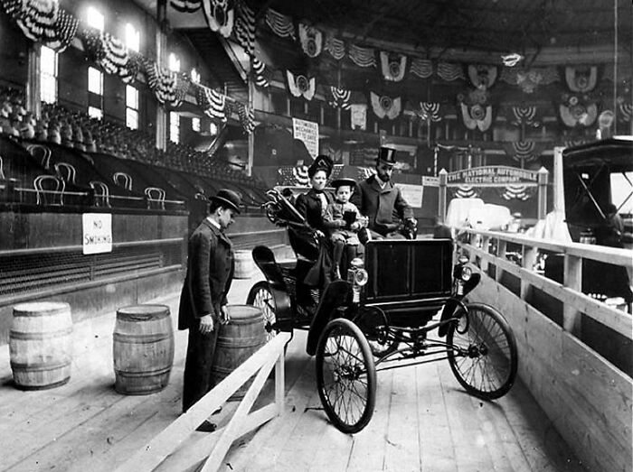 Early 1900s family riding a wild first car at an indoor exhibition showcasing the beginnings of automobile history.