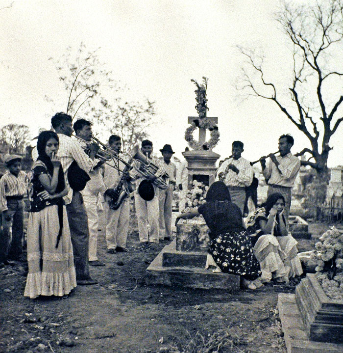 Black and white historical photo of people playing instruments and mourning at a decorated gravesite in a cemetery.