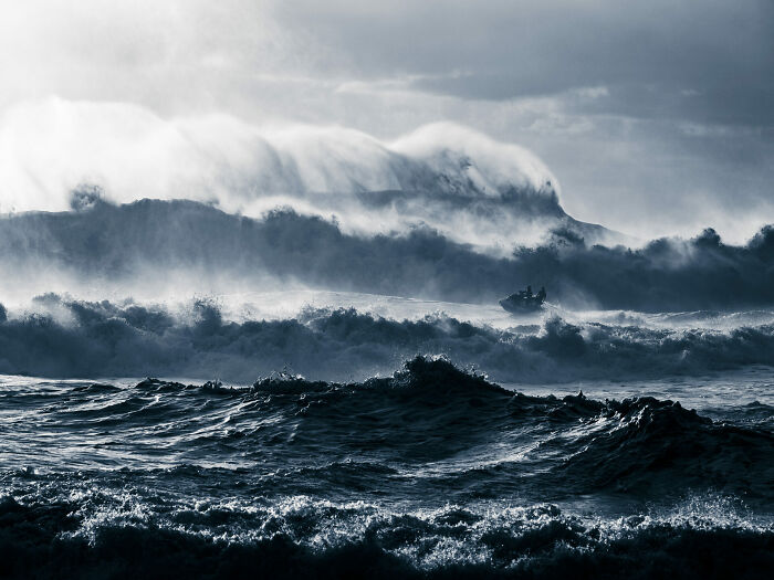 Rough ocean waves crashing with a small boat navigating through stormy waters in Ocean Photographer Of The Year Awards image.